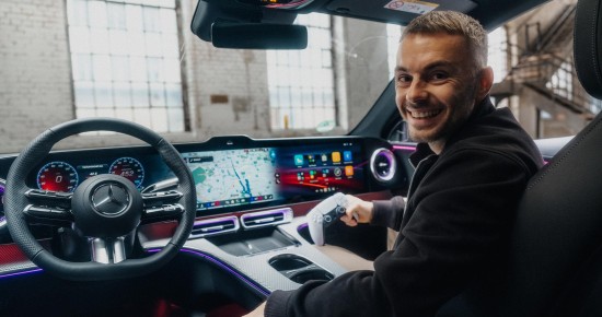 
					  Blick von der R�ckbank eines Mercedes-Benz auf Christopher Hartung von Mercedes-Benz, der auf dem Beifahrersitz sitzt, Kopfh�rer tr�gt und l�chelnd in die Kamera schaut, w�hrend er in der rechten Hand einen Gaming-Controller h�lt.
					  