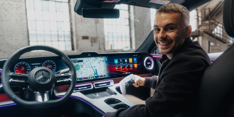 
				Blick von der R�ckbank eines Mercedes-Benz auf Christopher Hartung von Mercedes-Benz, der auf dem Beifahrersitz sitzt, Kopfh�rer tr�gt und l�chelnd in die Kamera schaut, w�hrend er in der rechten Hand einen Gaming-Controller h�lt.
				