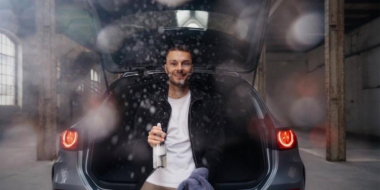 
				Frontaler Blick auf den ge�ffneten Kofferraum eines Mercedes-Benz GLC, in dem Christopher Hartung sitzt, in der linken Hand einen Putzlappen und in der rechten Hand eine Putzmittelspr�hflasche h�lt, mit der er in die Kamera spr�ht. Bekleidet ist Christoph mit einem wei�en T-Shirt und einer schwarzen Jacke mit Rei�verschluss.
				