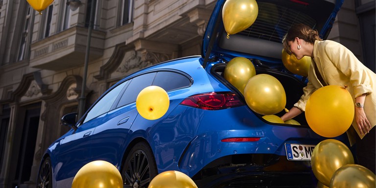 
				Eine Frau in einem gelben Blazer beugt sich in den ge�ffneten Kofferraum eines Mercedes-Benz CLA Shooting Brake in der Farbe klarblau metallic, aus dem goldene und gelbe Luftballons herausstr�men. Einige Ballons schweben bereits um das Fahrzeug herum, andere befinden sich noch im Kofferraum.
				