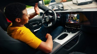 
Ein Mann im gelben T-Shirt sitzt auf dem Fahrersitz eines CLA und winkt in Richtung des Zentraldisplays, auf dem gerade ein Videocall mit einem Hund und deren Besitzerin stattfindet.
