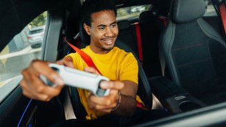 
Ein Mann im gelben T-Shirt sitzt auf dem Beifahrersitz eines CLA und h�lt l�chelnd einen Gaming-Controller in die Kamera.
