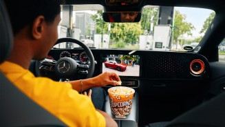 
Blick vom R�cksitz auf die Mittelkonsole, auf der ein gelber Popcorn-Eimer steht. Auf dem Fahrersitz sitzt ein Mann im gelben T-Shirt, der aus dem Eimer Popcorn isst, w�hrend auf dem Zentralbildschirm eine Folge &bdquo;Let&rsquo;s Drive Mercedes&ldquo; �ber den elektrischen CLA l�uft.
