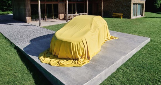 
					  Der neue GLA von Mercedes-Benz ist mit einem gelben Tuch verdeckt und steht auf dem Parkplatz vor einem modernen Wohnhaus auf einer gr�nen Wiese.
					  