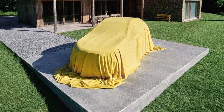 
				Der neue GLA von Mercedes-Benz ist mit einem gelben Tuch verdeckt und steht auf dem Parkplatz vor einem modernen Wohnhaus auf einer gr�nen Wiese.
				