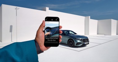 
					  Eine Hand h�lt ein Smartphone, dahinter ist ein silberner Mercedes-Benz vor wei�er, moderner Architektur und blauem Himmel zu sehen.
					  