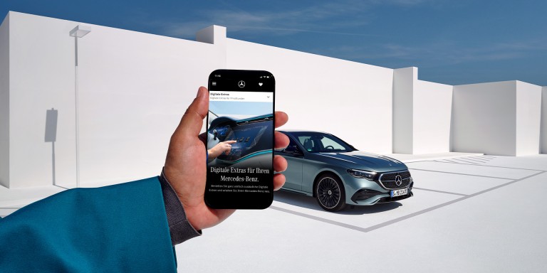 
				Eine Hand h�lt ein Smartphone, dahinter ist ein silberner Mercedes-Benz vor wei�er, moderner Architektur und blauem Himmel zu sehen.
				