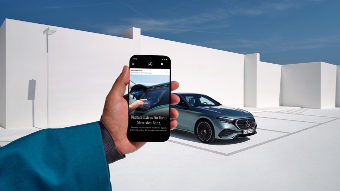 
				Eine Hand h�lt ein Smartphone, dahinter ist ein silberner Mercedes-Benz vor wei�er, moderner Architektur und blauem Himmel zu sehen.
				