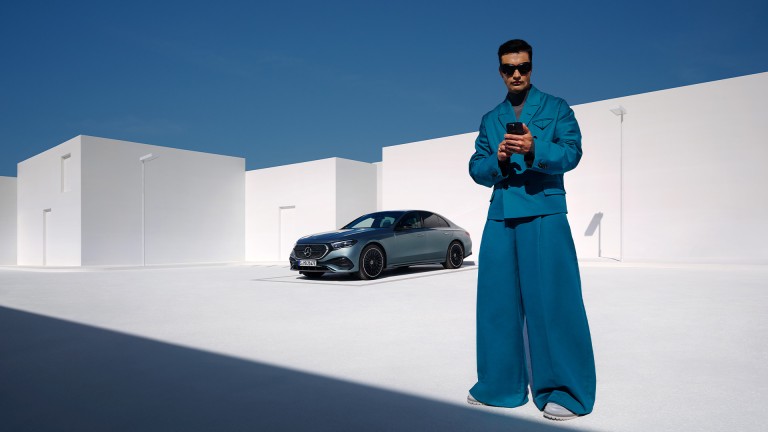 
			Ein Mann in blauem Anzug und mit Sonnenbrille h�lt ein Smartphone vor einem silbernen Mercedes-Benz und wei�er, moderner Architektur und blauem Himmel.
		