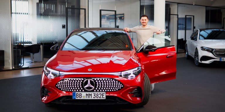 
				Felix B�llmann, Customer Onboarding f�r Digitale Extras bei Mercedes-Benz, steht l�chelnd neben der ge�ffneten Fahrert�r eines elektrischen Mercedes-Benz CLA in MANUFAKTUR patagonienrot metallic.
				