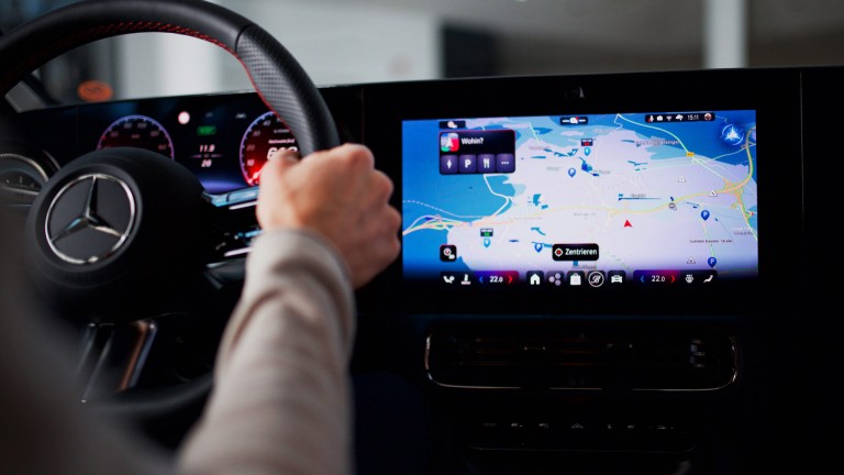 
			Blick von der R�ckbank auf den Zentralbildschirm eines CLA: Links ist die Hand des Fahrers am Lenkrad zu sehen, rechts der Zentralbildschirm mit der Navigationsansicht.
		