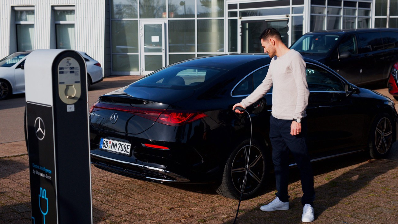 
			Felix B�llmann, Customer Onboarding f�r Digitale Extras bei Mercedes-Benz, steht vor einem Geb�ude neben einer Lades�ule und steckt das Ladekabel in die Buchse eines schwarzen elektrischen Mercedes-Benz CLA.
		