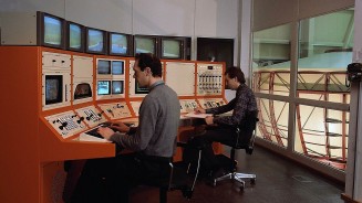 
Ein Bild aus 1985 zeigt zwei M�nner, die vor einem orange-wei�en Schaltpult mit Tasten und Bildschirmen sitzen, �ber das sie Daten des Simulators sammeln, der auf der rechten Seite des Bildes im Raum nebenan zu sehen ist..
