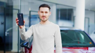 
Felix B�llmann steht vor einem roten CLA von Mercedes-Benz und h�lt mit seiner rechten Arm ein Smartphone in die Kamera.
