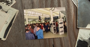 
					  Ein Bild von 1995 liegt neben anderen alten Fotos auf einem Holztisch. Darauf stehen zahlreiche Menschen dicht gedr�ngt in der damaligen Verkaufsstelle des Firmenangeh�rigengesch�fts in Sindelfingen. Ebenfalls zu sehen: Informationsst�nde, Aufsteller mit Fahrzeugabbildungen und Ausstattungsoptionen; im Hintergrund h�ngen Banner mit Modellhinweisen zu damals aktuellen E-Klasse Modellen.
					  