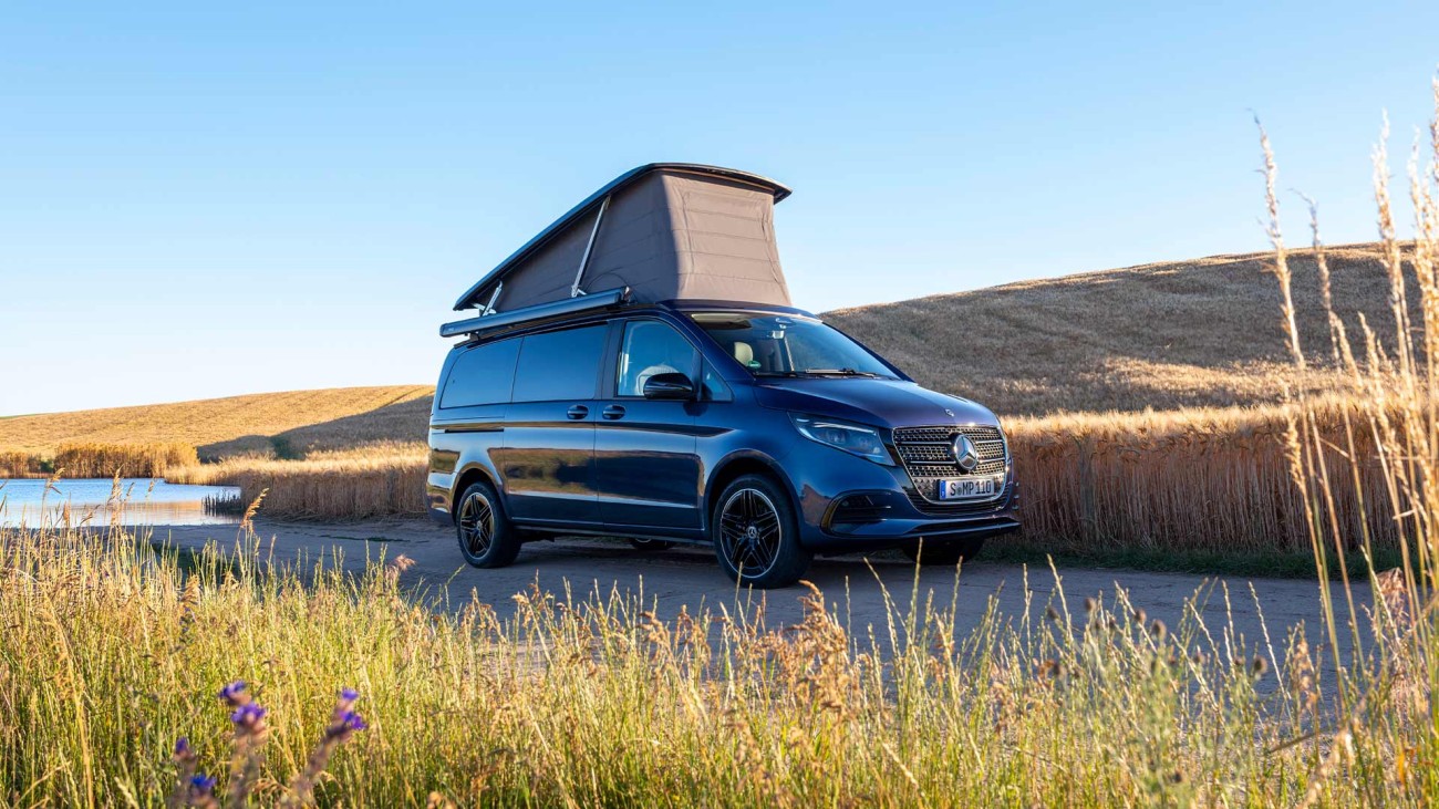 
			Ein Mercedes-Benz Marco Polo in sodalithblau metallic steht mit ge�ffnetem Aufstelldach auf einer von hohem Gras ges�umten Stra�e unter strahlend blauem Himmel, im Hintergrund ist links ein Gew�sser zu erkennen. Das Motiv ist von schr�g unten fotografiert.
		