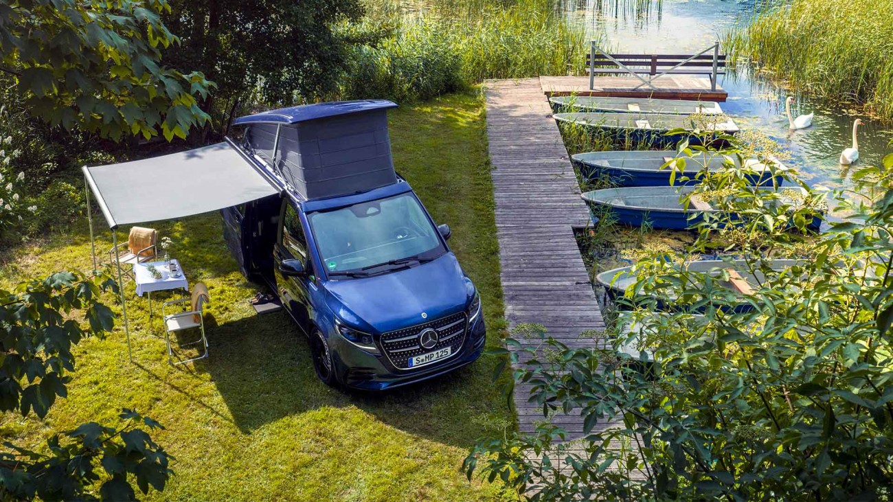 
			Ein Mercedes-Benz Marco Polo in sodalithblau metallic parkt an einem See links neben einem Holzsteg, an dem rechts Ruderboote befestigt sind. Das Aufstelldach und die Markise des MarcoPolo sind ausgefahren, vor der ge�ffneten Seitent�r stehen zwei Campingst�hle und ein Campingtisch Tisch. Das Motiv ist aus der Vogelperspektive fotografiert.
		