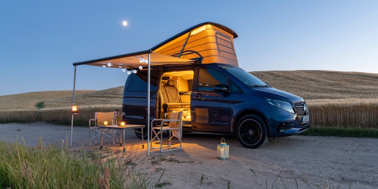 
				Seitliche Ansicht des neuen Mercedes-Benz Marco Polo in der Farbe sodalithblau metallic, der bei einsetzender D�mmerung in einer weitl�ufigen Landschaft steht. Das Aufstelldach ist ge�ffnet, die Markise ausgefahren und der beleuchtete Innenraum durch die ge�ffnete Schiebet�r sichtbar. Unter der Markise stehen ein Tisch und zwei Campingst�hle. Im Hintergrund erstrecken sich Felder und sanfte H�gel unter blauem Himmel.
				