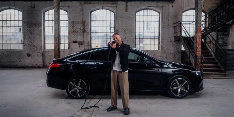 
				Christopher Hartung steht neben einem elektrischen CLA von Mercedes-Benz in nachtschwarz uni. In der Hand h�lt er das andere Ende des in das Fahrzeug eingesteckten Ladekabels und zielt spielerisch mit dem Stecker in die Kamera.
				