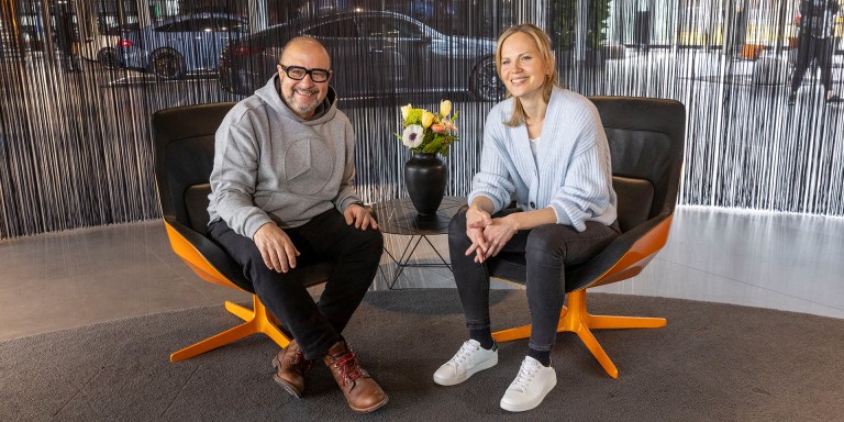 
				Frank Kemmerer sitzt links neben Veronika Kl�nder in einem modernen Sitzbereich im Mercedes-Benz Kundencenter Sindelfingen. Zwischen ihnen steht ein kleiner runder Tisch, darauf eine Vase mit Blumenstrau� in Gelb, Orange und Wei�. Beide l�cheln sitzend, leicht nach vorne gebeugt, in die Kamera. Frank tr�gt einen hellgrauen Kapuzenpullover mit Mercedes-Stern, eine dunkle Hose und braune Schuhe. Veronika tr�gt eine hellblaue Strickjacke, schwarze Jeans und wei�e Sneaker. Im Hintergrund ist ein dunkler Fadenvorhang zu sehen. Hinter dem Vorhang erkennt man schemenhaft Ausstellungsfahrzeuge.
				