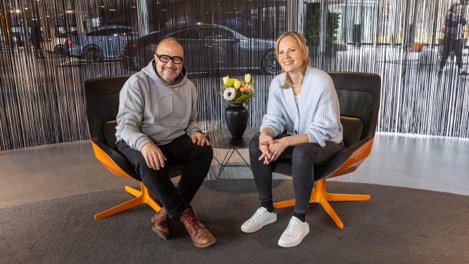 
				Frank Kemmerer sitzt links neben Veronika Kl�nder in einem modernen Sitzbereich im Mercedes-Benz Kundencenter Sindelfingen. Zwischen ihnen steht ein kleiner runder Tisch, darauf eine Vase mit Blumenstrau� in Gelb, Orange und Wei�. Beide l�cheln sitzend, leicht nach vorne gebeugt, in die Kamera. Frank tr�gt einen hellgrauen Kapuzenpullover mit Mercedes-Stern, eine dunkle Hose und braune Schuhe. Veronika tr�gt eine hellblaue Strickjacke, schwarze Jeans und wei�e Sneaker. Im Hintergrund ist ein dunkler Fadenvorhang zu sehen. Hinter dem Vorhang erkennt man schemenhaft Ausstellungsfahrzeuge.
				