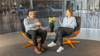 
Frank Kemmerer sitzt links neben Veronika Kl�nder in einem modernen Sitzbereich im Mercedes-Benz Kundencenter Sindelfingen. Zwischen ihnen steht ein kleiner runder Tisch, darauf eine Vase mit Blumenstrau� in Gelb, Orange und Wei�. Sie unterhalten sich und gestikulieren. Frank tr�gt einen hellgrauen Kapuzenpullover mit Mercedes-Stern, eine dunkle Hose und braune Schuhe. Veronika tr�gt eine hellblaue Strickjacke, schwarze Jeans und wei�e Sneaker. Im Hintergrund ist ein dunkler Fadenvorhang zu sehen. Hinter dem Vorhang erkennt man schemenhaft Ausstellungsfahrzeuge.
