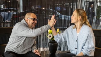 
Frank Kemmerer sitzt links neben Veronika Kl�nder in einem modernen Sitzbereich im Mercedes-Benz Kundencenter Sindelfingen. Zwischen ihnen steht ein kleiner runder Tisch, darauf eine Vase mit Blumenstrau� in Gelb, Orange und Wei�. Die beiden klatschen mit der rechten Hand ab. Frank tr�gt einen hellgrauen Kapuzenpullover mit Mercedes-Stern, eine dunkle Hose und braune Schuhe. Veronika tr�gt eine hellblaue Strickjacke, schwarze Jeans und wei�e Sneaker. Im Hintergrund ist ein dunkler Fadenvorhang zu sehen. Hinter dem Vorhang erkennt man schemenhaft Ausstellungsfahrzeuge.
