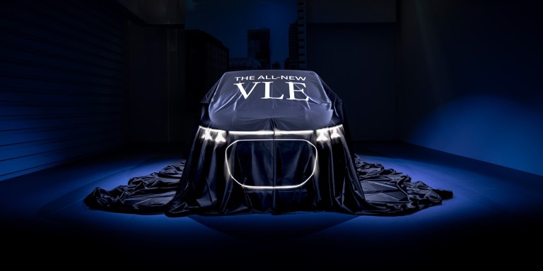 
				Ein unter einem dunklen Tuch verborgenes Fahrzeug steht auf einer B�hne in blauem Licht. Auf der Abdeckung steht &bdquo;The all-new VLE&ldquo;. Die Silhouette, die Front und die leuchtenden Scheinwerfer sind bereits erkennbar.
				