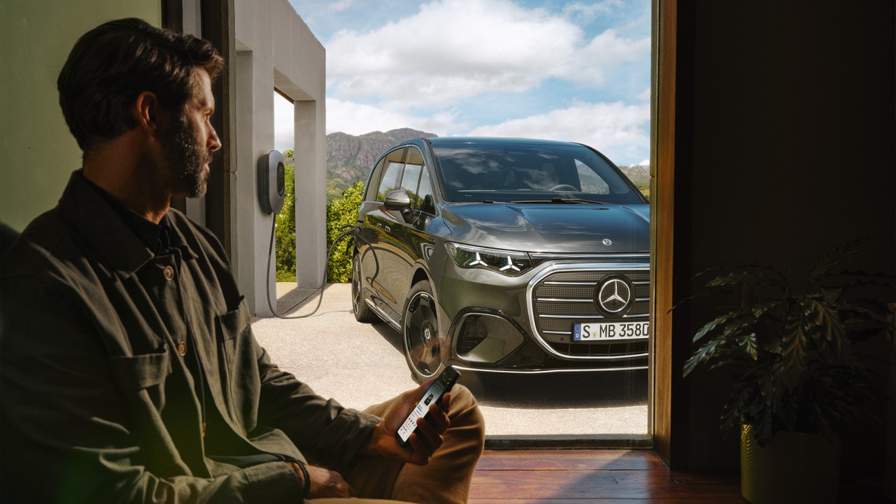 
			Im Hintergrund ist der Mercedes-Benz VLE zu sehen, der an einer Ladestation aufgeladen wird. Im Vordergrund sitzt ein Mann, der auf seinem Smartphone den Ladevorgang verfolgt.
		
