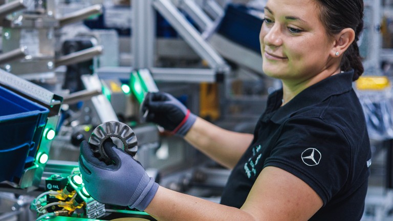 
			Mitarbeiterin in der Produktion h�lt ein Metallbauteil in der Hand und pr�ft es an einer industriellen Fertigungsanlage.
		