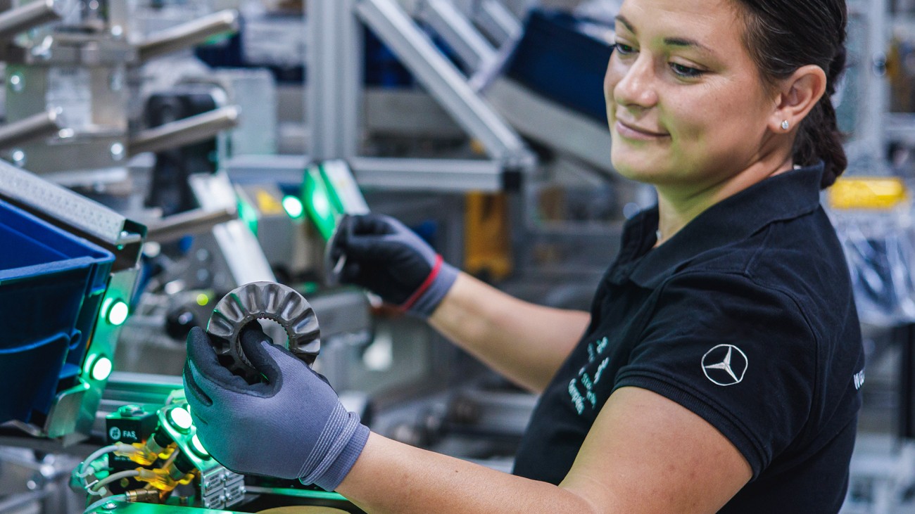 
			Mitarbeiterin in der Produktion h�lt ein Metallbauteil in der Hand und pr�ft es an einer industriellen Fertigungsanlage.
		