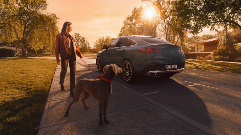 
			Seitliche R�ckansicht des Mercedes-Benz GLE Coup� auf einer ruhigen Wohnstra�e: Im Vordergrund steht eine Frau mit Hund, die auf das Fahrzeug blickt. Im Fokus stehen die flie�ende Coup�-Linie und das markante Heck.
		