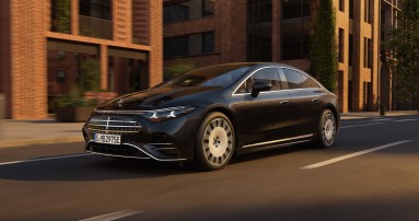 
					  Ein Mercedes-Benz EQS in MANUFAKTUR schwarz sparkling f�hrt in der Abendsonne auf einer Stra�e an einem Backsteingeb�ude mit gro�er Fensterfront vorbei.
					  