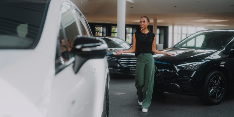 
				Mercedes-Benz Moderatorin Yasmine Blair geht lchelnd durch eine helle Austellungshalle zwischen mehreren Mercedes-Benz Fahrzeugen.
				