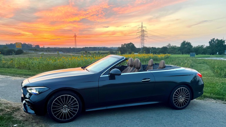 
			Irinas schwarzes CLE Cabriolet parkt auf einem Feldweg, umrahmt von grnen Wiesen und Bumen, im Hintergrund ist ein sommerlicher Sonnenuntergang zu sehen.
		