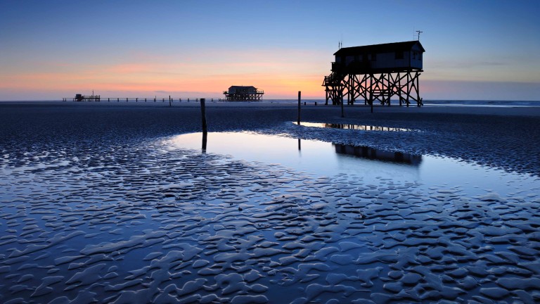 
			Besinnlicher Sonnenuntergang in St. Peter-Ording, Blick auf die typischen Pfahlhuser entlang des Sandstrands.
		