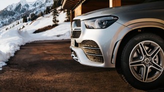 
Seitenansicht eines silbernen Mercedes-Benz, der auf einem vom Schnee befreiten Parkplatz parkt, im Hintergrund sind verschneite Berge, Hnge und Tannenbume zu sehen.
