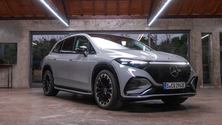 
			Der EQS SUV von Mercedes-Benz in diamantweiß metallic BRIGHT steht in einer Halle vor einer geöffneten Einfahrt.
		