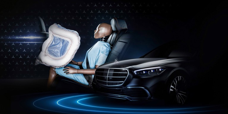 
				Links ist ein Dummy in blauer Kleidung auf einem Fondsitz zu sehen, vor ihm ein entfalteter Frontalairbag, rechts daneben die Motorhaube eines schwarzen Mercedes-Benz Fahrzeugs mit Stern auf der Haube.
				