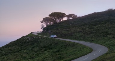 
						  Ein EQE von Mercedes-Benz in hightechsilber fhrt auf einer kurvigen Strae, umgeben von Bumen und bewachsenen Berghngen.
						  