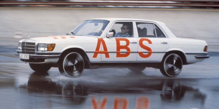 
				Eine S-Klasse Limousine der Baureihe 116 fhrt ber eine nasse Strae, ohne dass die Rder blockieren. Auf der linken Fahrzeugseite und der Motorhaube stehen in Rot die Buchstaben „ABS“, am Steuer sitzt ein Mann mit Sonnenbrille. Bildquelle: Mercedes-Benz Classic.
				
