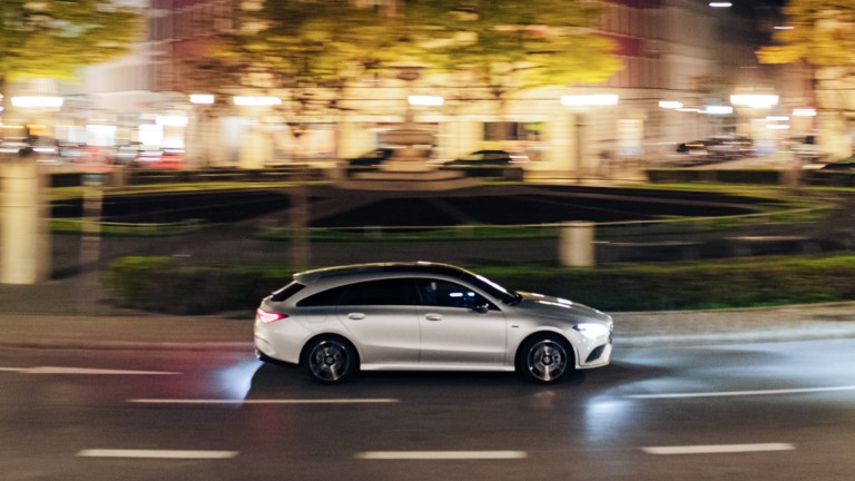 
			Ein CLA Shooting Brake in hightechsilber metallic fhrt entlang eines Kreisverkehrs, im Hintergrund sind verschwommen die abendlich leuchtenden Straenlaternen, Bume und einige Huser einer Stadt zu sehen.
		