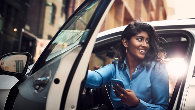 
			Eine Frau in einer blauen Bluse steigt aus einem Mercedes-Benz Fahrzeug aus und hlt dabei ihr Smartphone in der Hand.
		