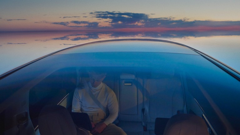 
			Blick von auen durch das Panoramadach eines  
EQS SUV von Mercedes-Benz. Eine Person sitzt auf der Rckbank, im Hintergrund ist der rtliche Himmel beim Sonnenuntergang zu sehen.
		