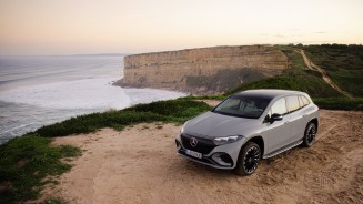 
Ein Mercedes-Benz EQS steht auf sandigem Untergrund, im Hintergrund ist das Meer und eine grn bewachsene Klippe zu sehen.
