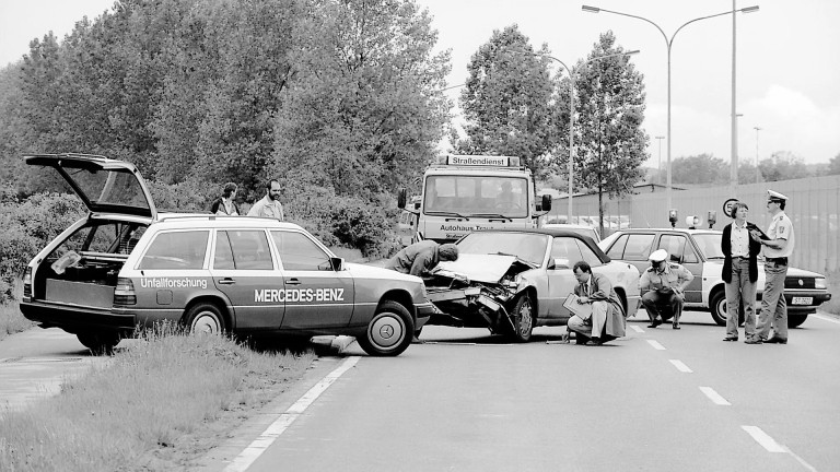 
			Eine Schwarz-Wei-Aufnahme einer Unfallszene im Jahr 1969: Links am Straenrand steht ein Mercedes-Benz Polizeifahrzeug mit geffneter Heckklappe, rechts daneben stehen fnf Menschen, die das in der Mitte der Fahrbahn stehende Unfallfahrzeug untersuchen.
		