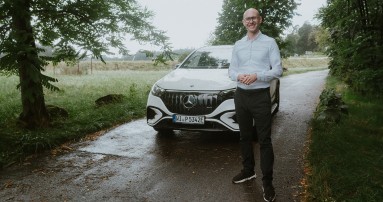 
						  Mercedes-Benz Produkttrainer Christoph Geissler steht vor einem Mercedes-AMG EQE 43 SUV in MANUFAKTUR opalithwei bright, der auf einer von Bumen gesumten Landstrae parkt.
						  