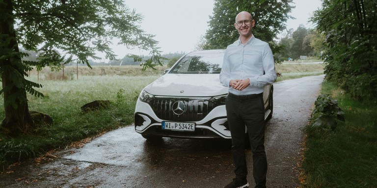 
				Mercedes-Benz Produkttrainer Christoph Geissler steht vor einem Mercedes-AMG EQE 43 SUV in MANUFAKTUR opalithwei bright, der auf einer von Bumen gesumten Landstrae parkt.
				