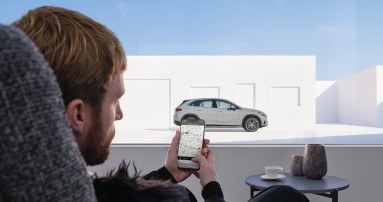 
						  Ein Mann sitzt an einem Tisch und schaut auf sein Handy, ber das er mit seinem Fahrzeug verbunden ist. Dieses ist durch das Fenster zu sehen.
						  