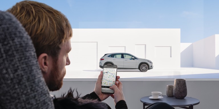 
				Ein Mann sitzt an einem Tisch und schaut auf sein Handy, ber das er mit seinem Fahrzeug verbunden ist. Dieses ist durch das Fenster zu sehen.
				